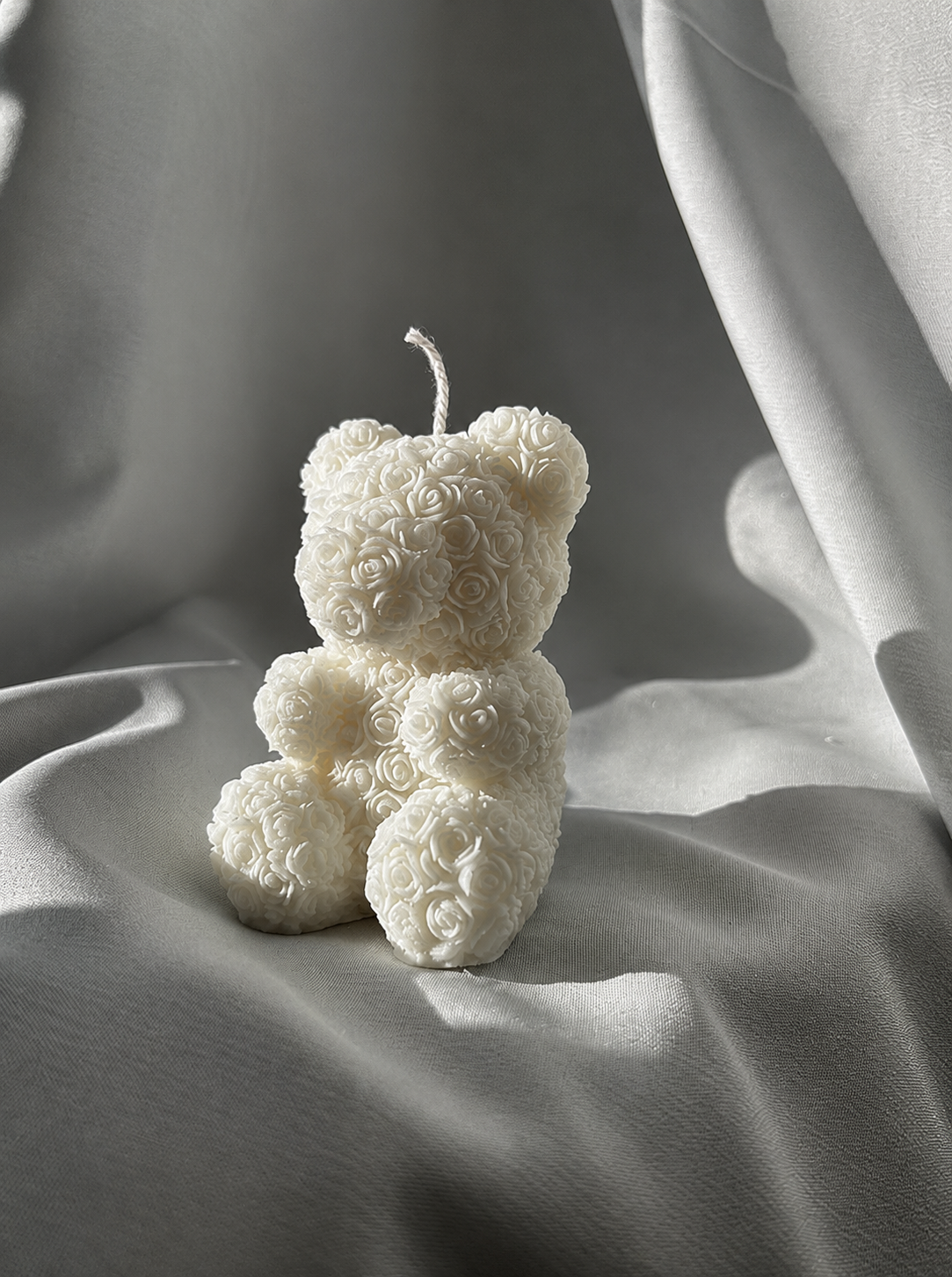 A small white aromatherapy candle designed to resemble a teddy bear with a floral design pattern on a textured gray surface 
